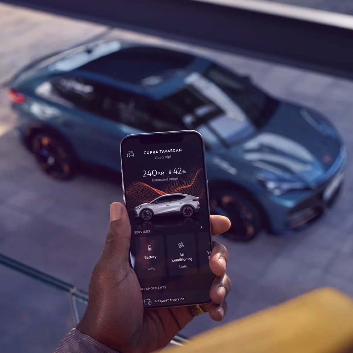 Pessoa a segurar um smartphone com a app My CUPRA visível. Ao fundo, CUPRA Tavascan VZ, ideal para renting empresarial.