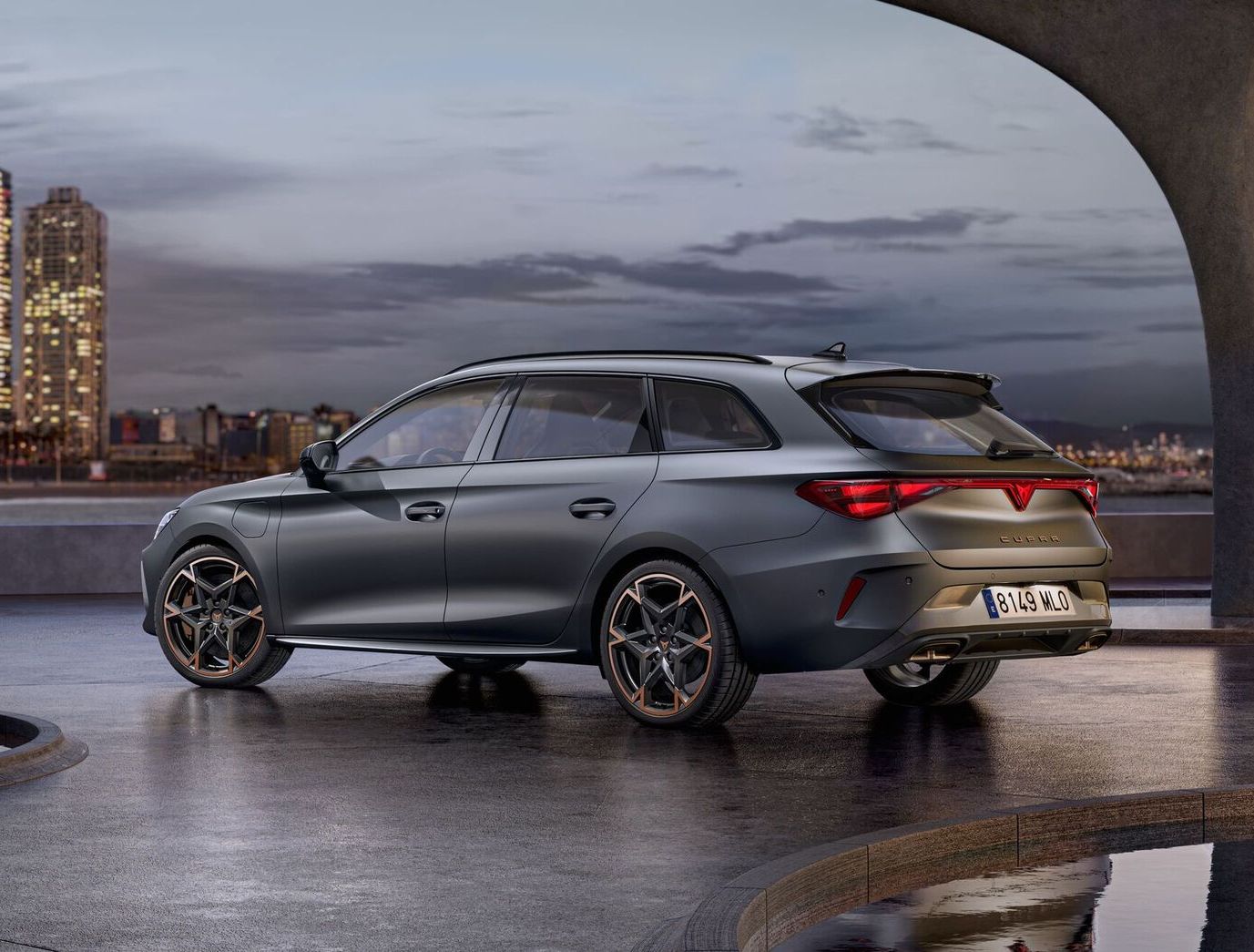 Vista lateral traseira do novo automóvel desportivo e aerodinâmico CUPRA Leon Sportstourer 2025 em cinzento mate, estacionado num passeio durante o anoitecer. A linha do horizonte da cidade brilha ao fundo.