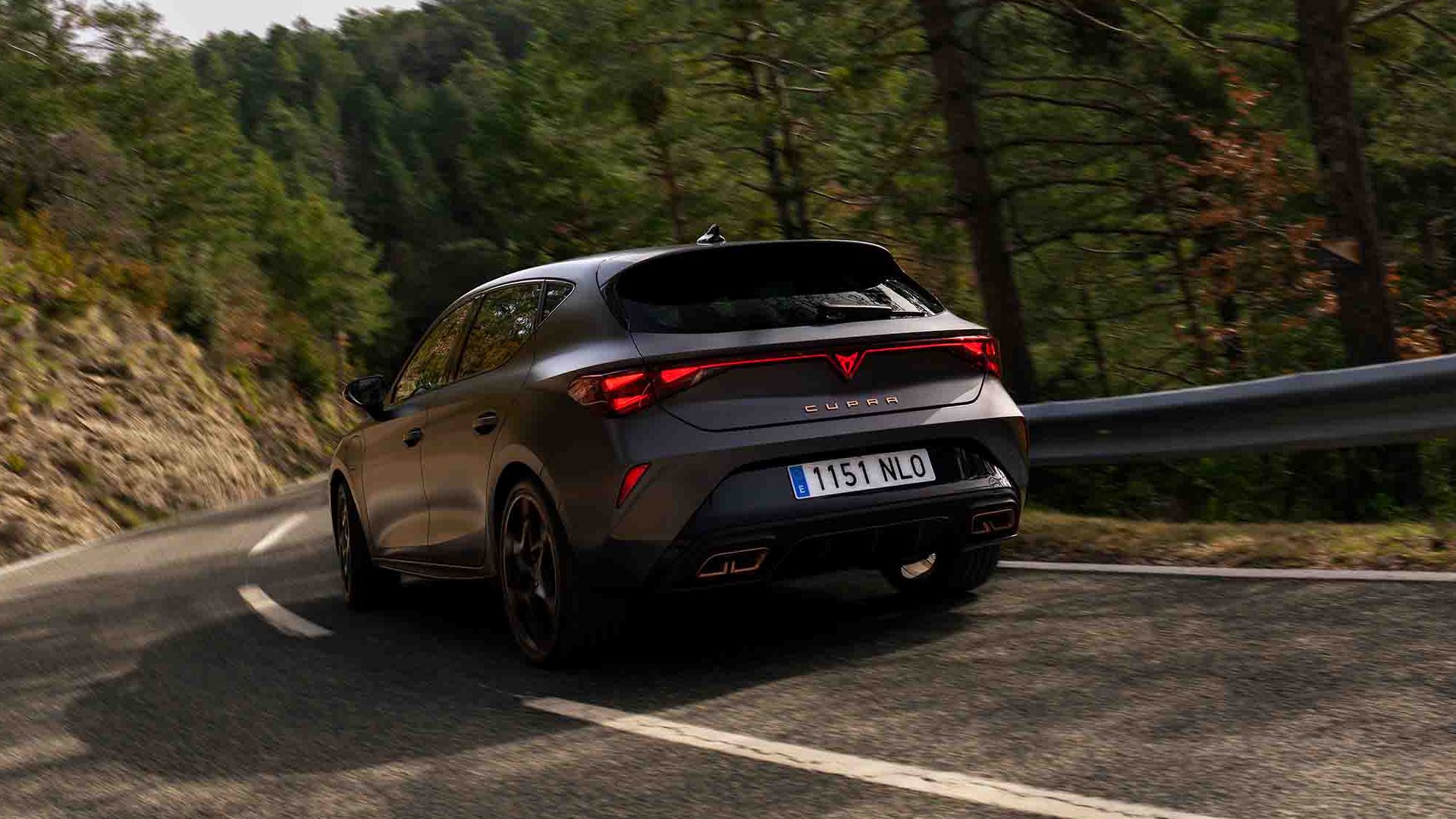 Novo CUPRA Leon PHEV em cinzento mate, com jantes forjadas em preto e cobre, iluminação traseira LED acesa, estacionado sobre piso em cimento junto à estrutura de rebordos brilhantes 