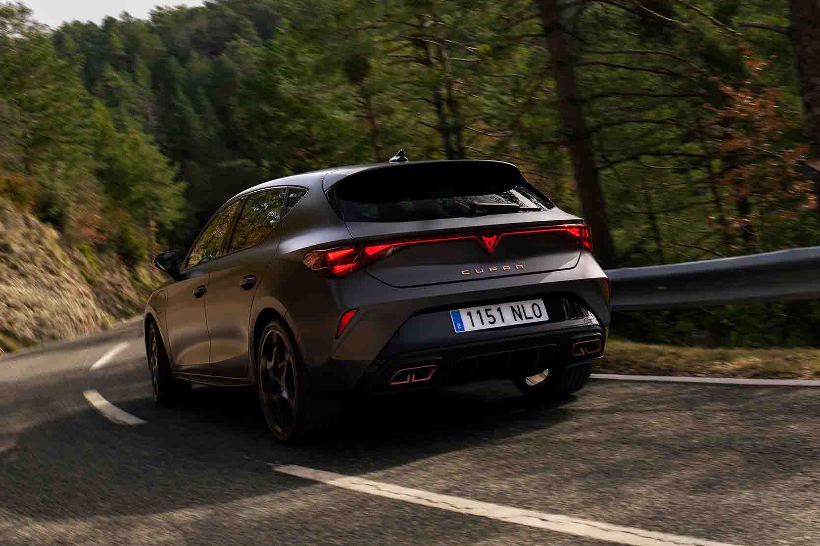 Novo CUPRA Leon PHEV em cinzento mate, com jantes forjadas em preto e cobre, iluminação traseira LED acesa, estacionado sobre piso em cimento junto à estrutura de rebordos brilhantes 