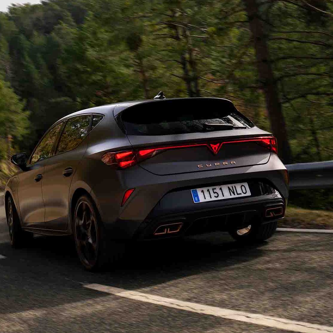 Novo CUPRA Leon PHEV em cinzento mate, com jantes forjadas em preto e cobre, iluminação traseira LED acesa, estacionado sobre piso em cimento junto à estrutura de rebordos brilhantes 