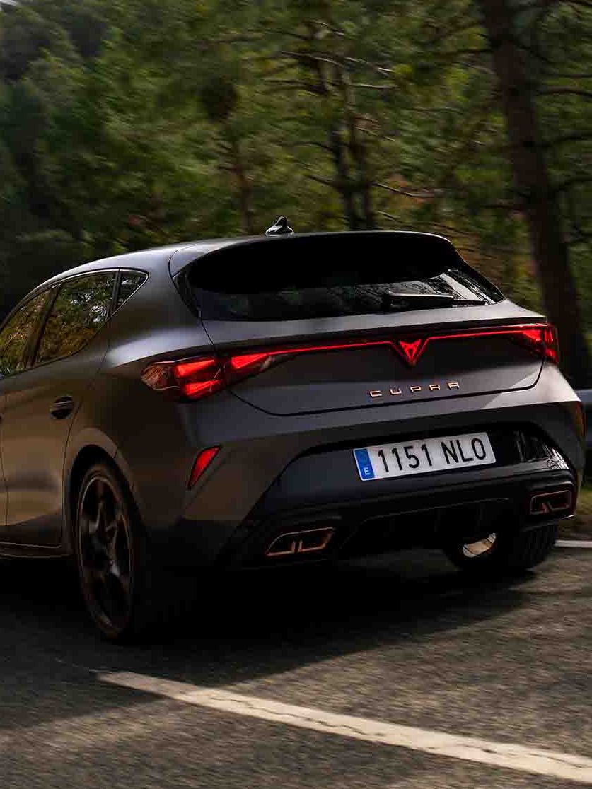Novo CUPRA Leon PHEV em cinzento mate, com jantes forjadas em preto e cobre, iluminação traseira LED acesa, estacionado sobre piso em cimento junto à estrutura de rebordos brilhantes 