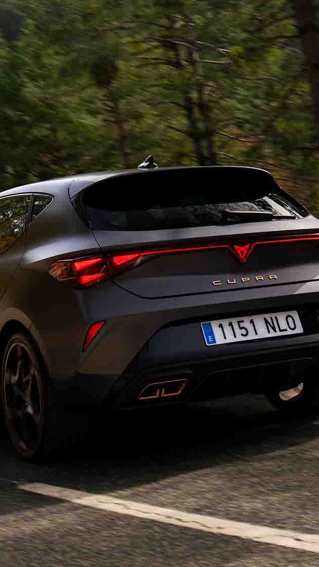 Novo CUPRA Leon PHEV em cinzento mate, com jantes forjadas em preto e cobre, iluminação traseira LED acesa, estacionado sobre piso em cimento junto à estrutura de rebordos brilhantes 
