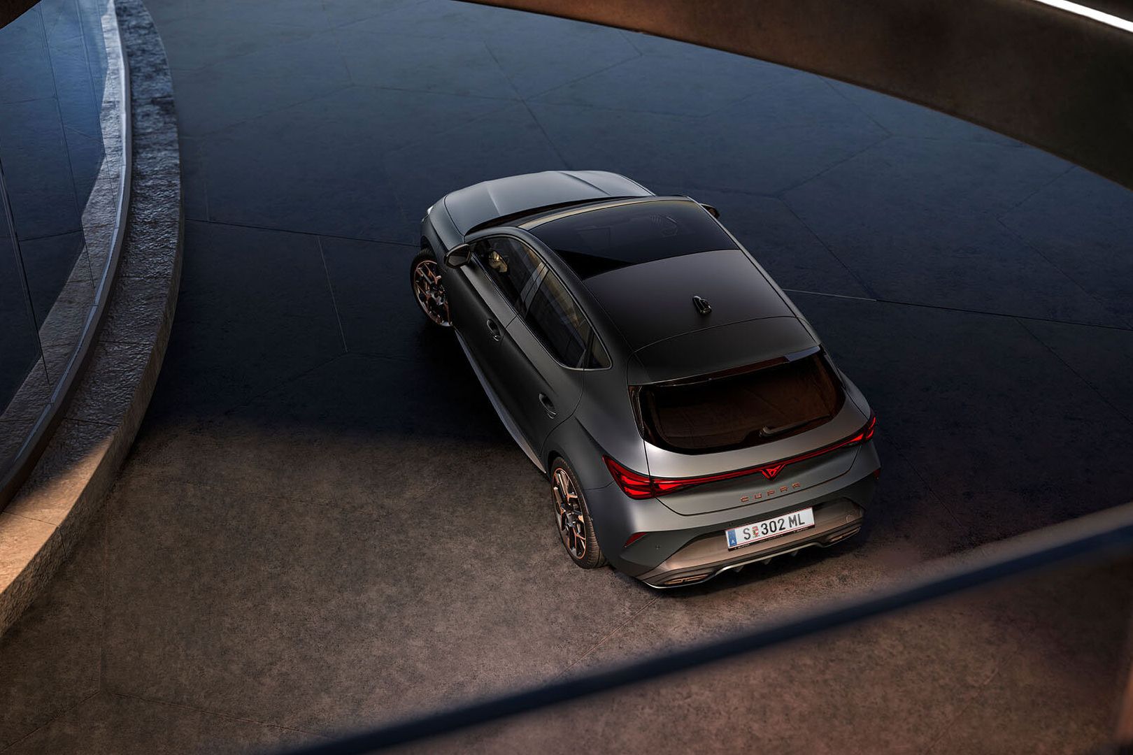 Novo CUPRA Leon 2025 PHEV em cinzento mate, com jantes forjadas em preto e cobre, iluminação traseira LED acesa, estacionado sobre piso em cimento junto a estrutura de rebordos brilhantes 