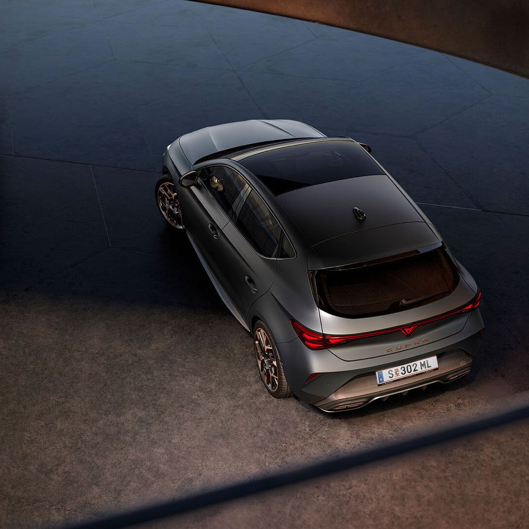 Novo CUPRA Leon 2025 PHEV em cinzento mate, com jantes forjadas em preto e cobre, iluminação traseira LED acesa, estacionado sobre piso em cimento junto a estrutura de rebordos brilhantes 