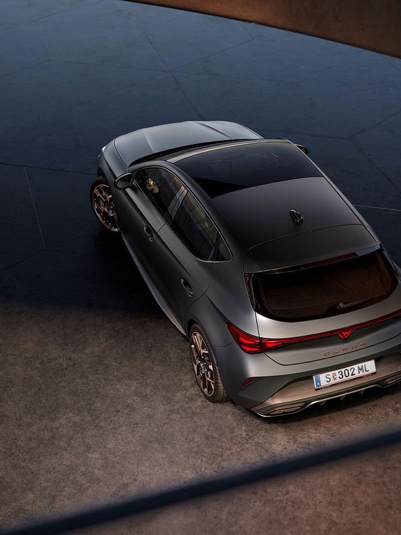 Novo CUPRA Leon 2025 PHEV em cinzento mate, com jantes forjadas em preto e cobre, iluminação traseira LED acesa, estacionado sobre piso em cimento junto a estrutura de rebordos brilhantes 