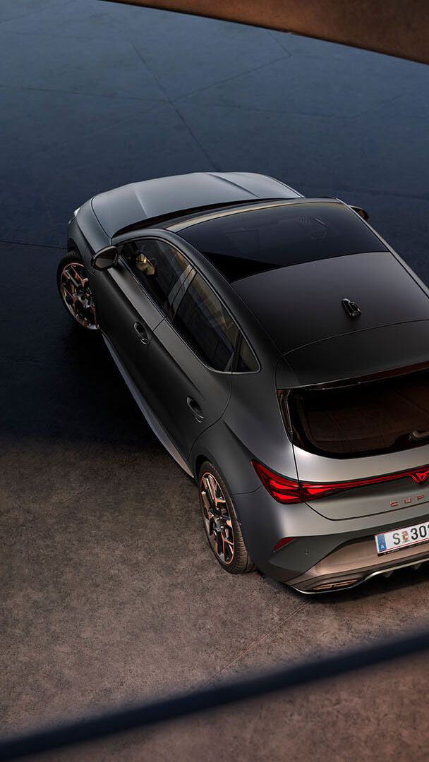 Novo CUPRA Leon 2025 PHEV em cinzento mate, com jantes forjadas em preto e cobre, iluminação traseira LED acesa, estacionado sobre piso em cimento junto a estrutura de rebordos brilhantes 