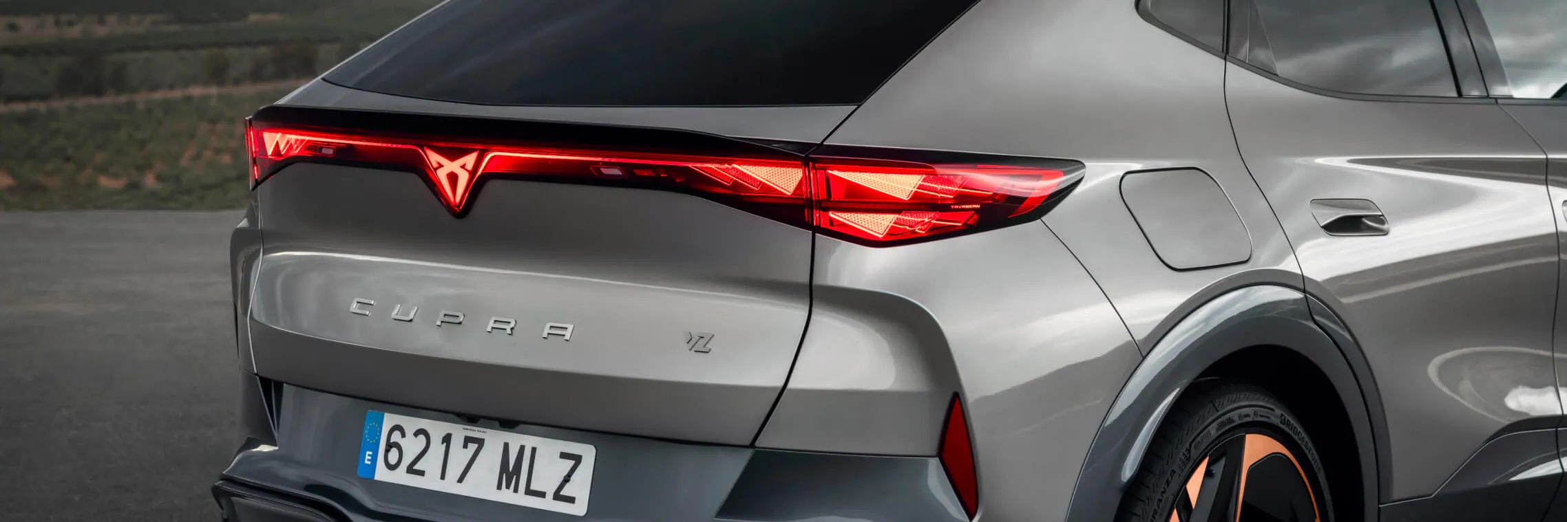 Vista traseira do CUPRA Tavascan cinzento, viatura de empresa, com jantes em cobre e luz contínua coast-to-coast.[