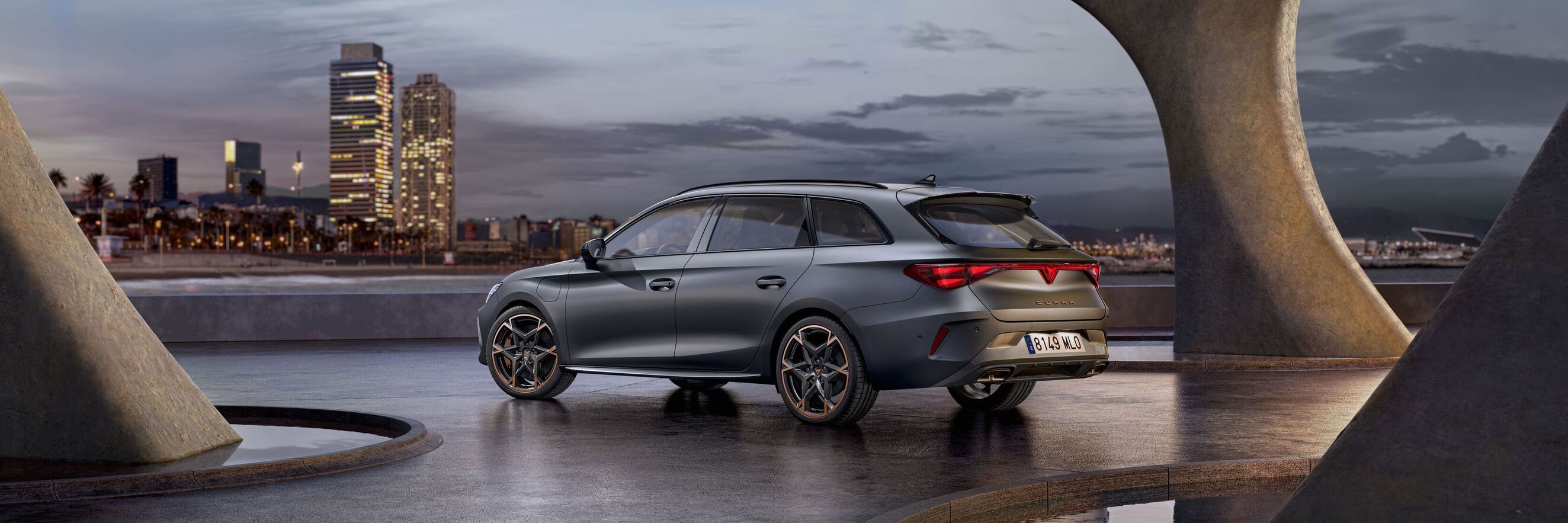 Vista lateral traseira do novo automóvel desportivo e aerodinâmico CUPRA Leon Sportstourer 2025 em cinzento mate, estacionado num passeio durante o anoitecer. A linha do horizonte da cidade brilha ao fundo.