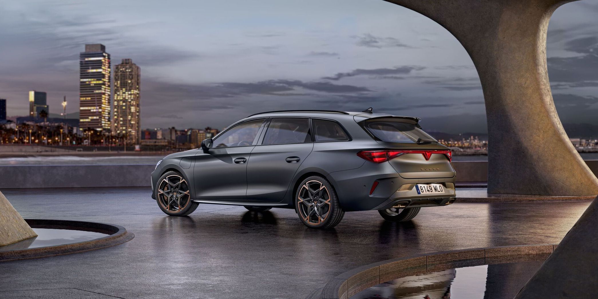 Vista lateral traseira do novo automóvel desportivo e aerodinâmico CUPRA Leon Sportstourer 2025 em cinzento mate, estacionado num passeio durante o anoitecer. A linha do horizonte da cidade brilha ao fundo.