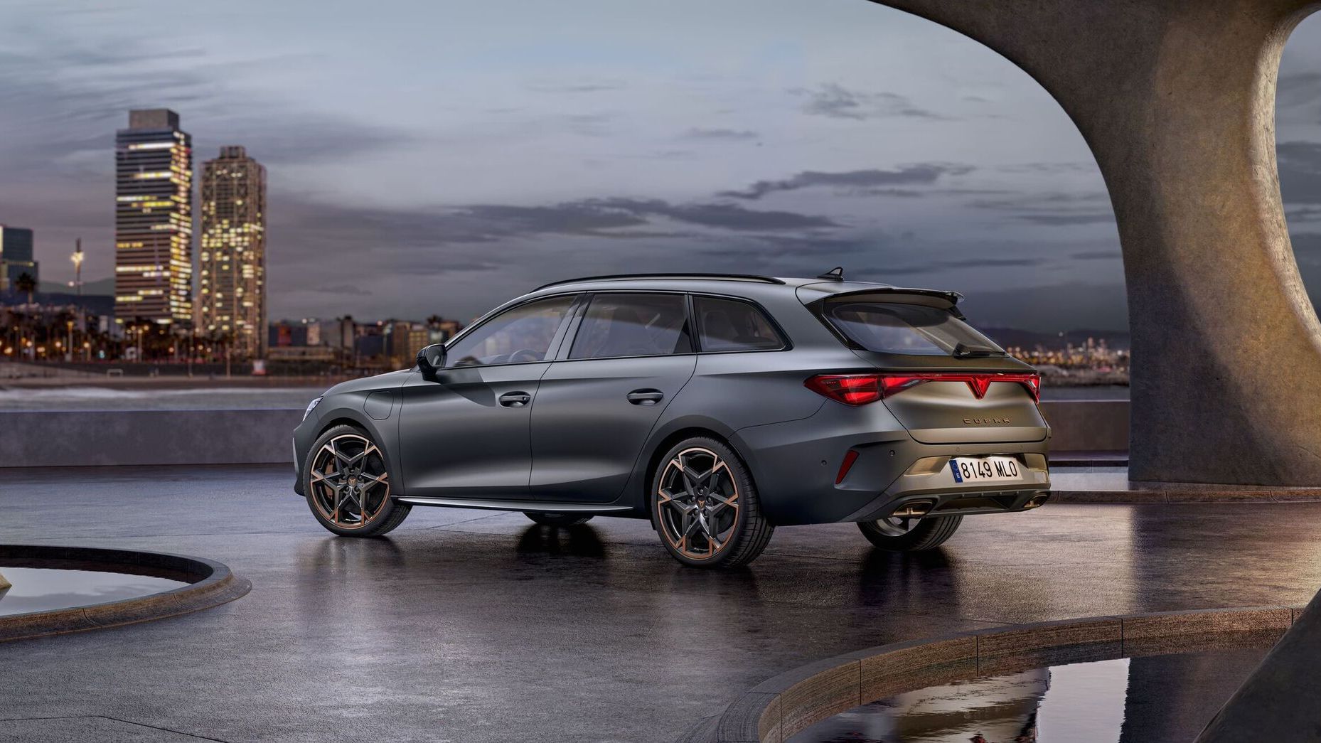 Vista lateral traseira do novo automóvel desportivo e aerodinâmico CUPRA Leon Sportstourer 2025 em cinzento mate, estacionado num passeio durante o anoitecer. A linha do horizonte da cidade brilha ao fundo.