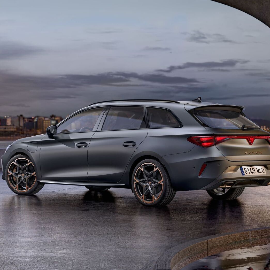 Vista lateral traseira do novo automóvel desportivo e aerodinâmico CUPRA Leon Sportstourer 2025 em cinzento mate, estacionado num passeio durante o anoitecer. A linha do horizonte da cidade brilha ao fundo.